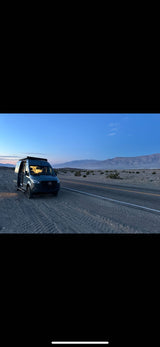 Mercedes Sprinter RV rental van, side door open, parked on a desert road shoulder at dusk with mountains in background.