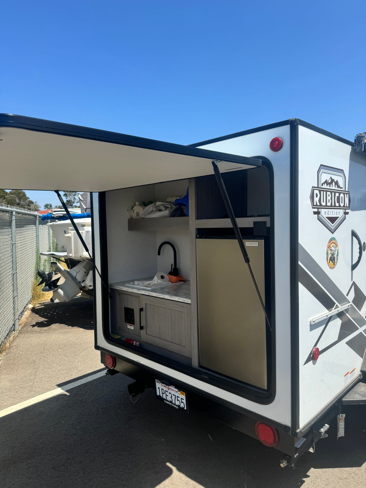 White RV rental with its rear hatch open, revealing an outdoor kitchen setup with a sink, faucet, and fridge. Parked under clear skies.