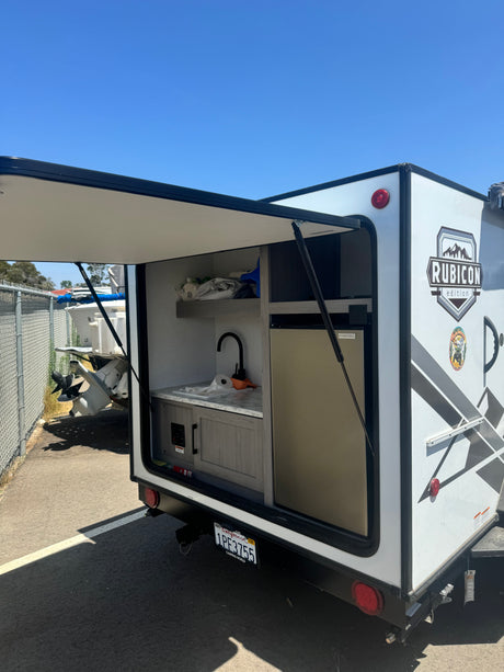 White RV rental with its rear hatch open, revealing an outdoor kitchen setup with a sink, faucet, and fridge. Parked under clear skies.