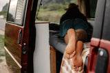 A person's bare feet are seen relaxing inside an RV rental, looking out at a scenic road and landscape. Perfect van life.