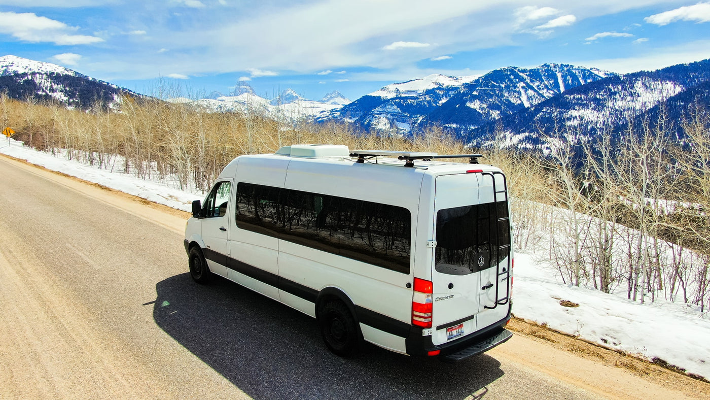 A white Sprinter RV rental parked on a mountain road with snow-covered peaks and bare trees under a blue sky. — photo 9