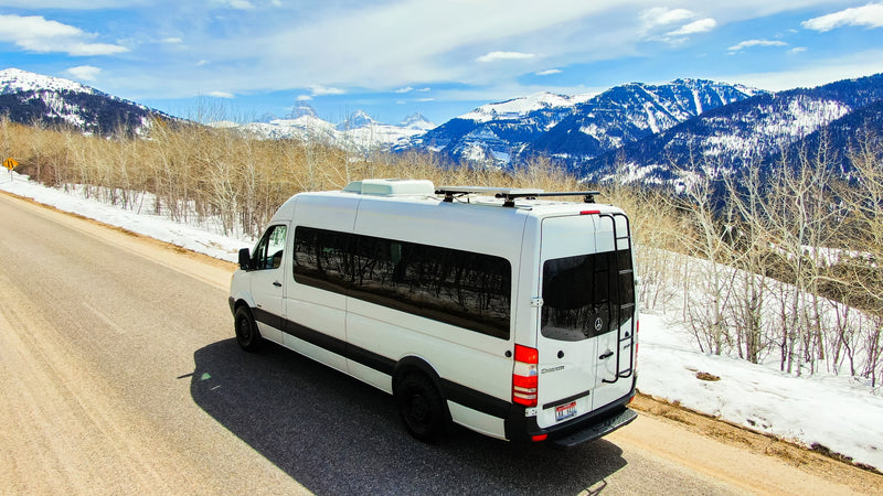 A white Sprinter RV rental parked on a mountain road with snow-covered peaks and bare trees under a blue sky. — photo 9