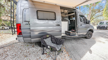 A gray Winnebago RV rental camper van with an extended awning, outdoor grill, and camping chair setup for adventure.