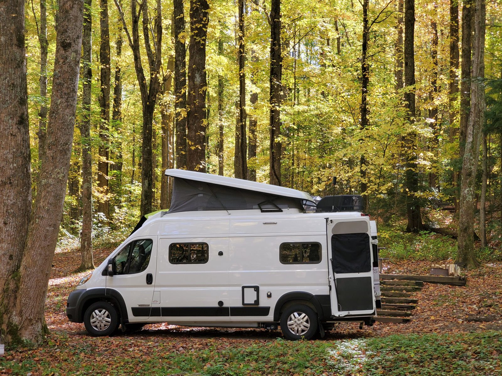 A white camper van (RV rental) with its pop-top roof open, parked in a vibrant autumn forest.