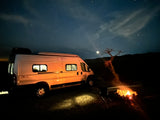 An RV rental camper van parked at night, illuminated by a glowing campfire. Stars twinkle above a gnarled tree and dark mountains.