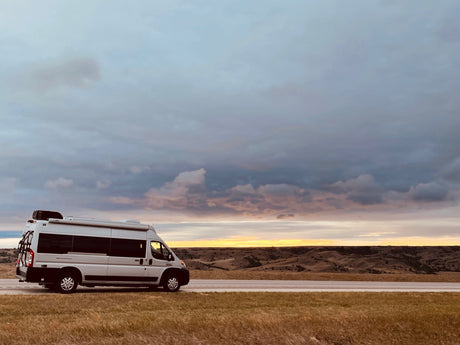 White RV rental on a roadside at dramatic sunset. Golden light illuminates rolling hills and expressive clouds.