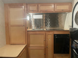 Clean RV rental kitchen with light wood cabinets, stainless steel backsplash, sink, cooktop, and compact fridge.