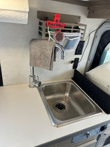 RV rental compact kitchenette: stainless sink, modern faucet, counter, & wall storage with "Prevent Wildfires!" clip.