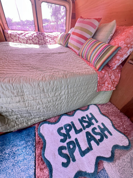 Relaxing RV rental interior featuring a bed with green comforter, colorful pillows, and a "Splish Splash" rug. Outside view.