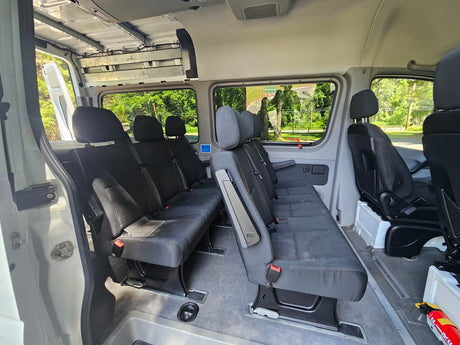 Spacious RV rental passenger van interior. Two rows of black bench seats, large windows, and an open side door for easy access.