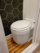 Compact RV rental bathroom features a white toilet, striking black hexagonal wall tiles, and a wooden slatted floor mat.
