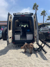 Custom RV rental camper van with open rear doors, a dog, and gear, parked on a sandy beach.