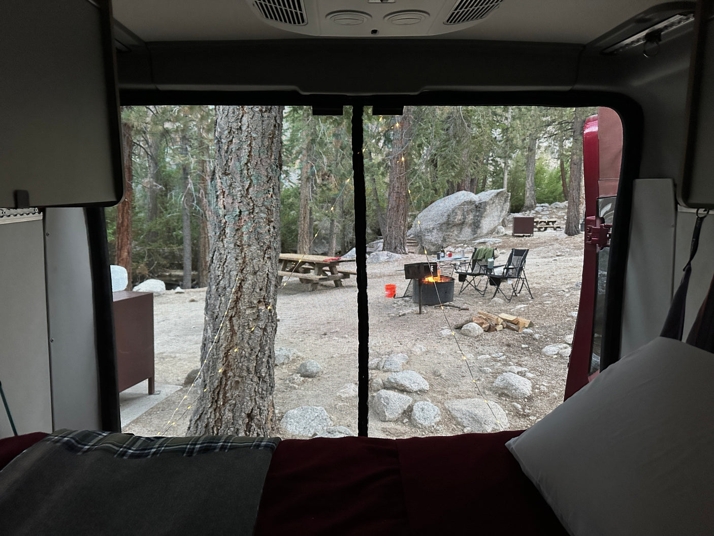 Cozy view from an RV rental, looking out at a peaceful campsite with a warm campfire, trees, and rocks. Perfect camping!