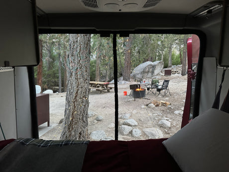 Cozy view from an RV rental, looking out at a peaceful campsite with a warm campfire, trees, and rocks. Perfect camping!