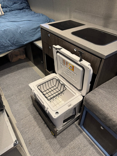 Inside an RV rental: compact kitchen with cooktop/sink, cozy bed, and an open Yeti cooler on a slide-out.