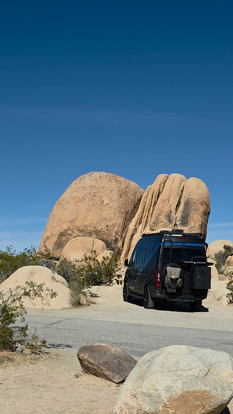 Black RV rental van parked in a sunny desert landscape with massive rock formations under a clear blue sky, seen from the rear.