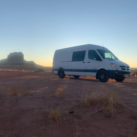 An RV rental, a white camper van, is parked at sunset in a vast desert with a distant mesa. Adventure awaits!