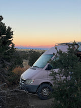 Light-colored RV rental campervan nestled in trees, with stunning sunrise views over distant snowy mountains. Adventure awaits!
