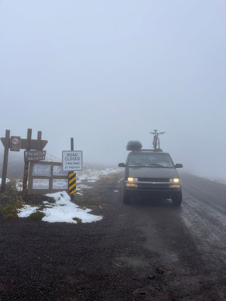 An rv rental van with bike & cargo stops at 'ROAD CLOSED' signs on a foggy, snowy road. Low visibility.