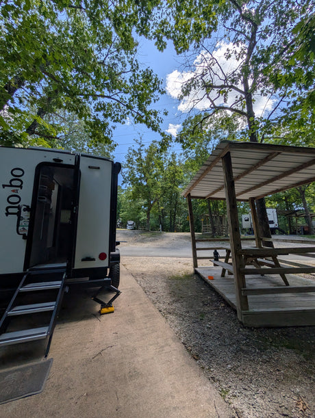 An RV rental campsite features a white travel trailer with steps down, next to a covered picnic area under green trees, ready for the outdoors.