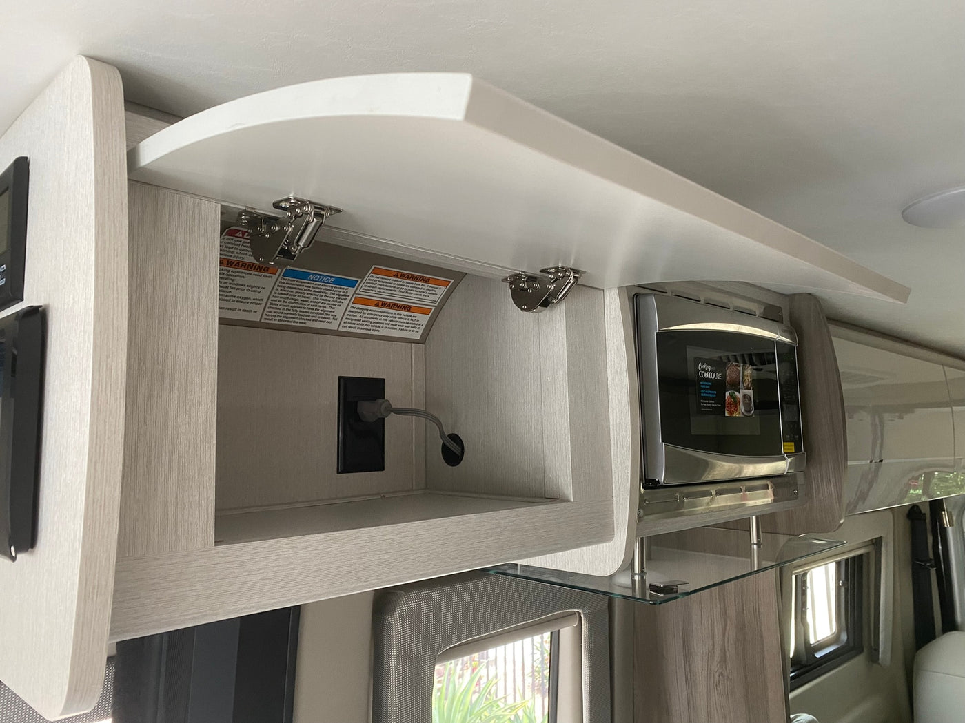 RV rental interior: open overhead cabinet with warning labels, power outlet, and a microwave oven. — photo 12