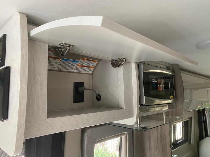 RV rental interior: open overhead cabinet with warning labels, power outlet, and a microwave oven. — photo 12