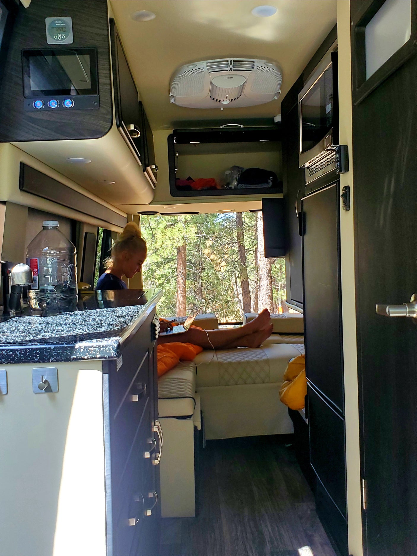 Person working on laptop inside a well-equipped RV rental, featuring a kitchen, microwave, and scenic forest views.
