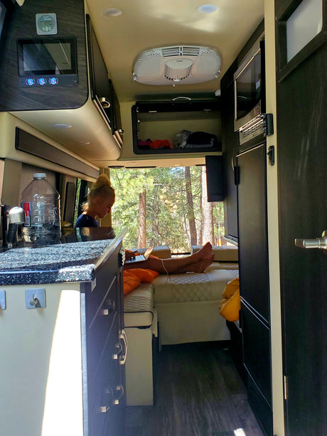 Person working on laptop inside a well-equipped RV rental, featuring a kitchen, microwave, and scenic forest views.
