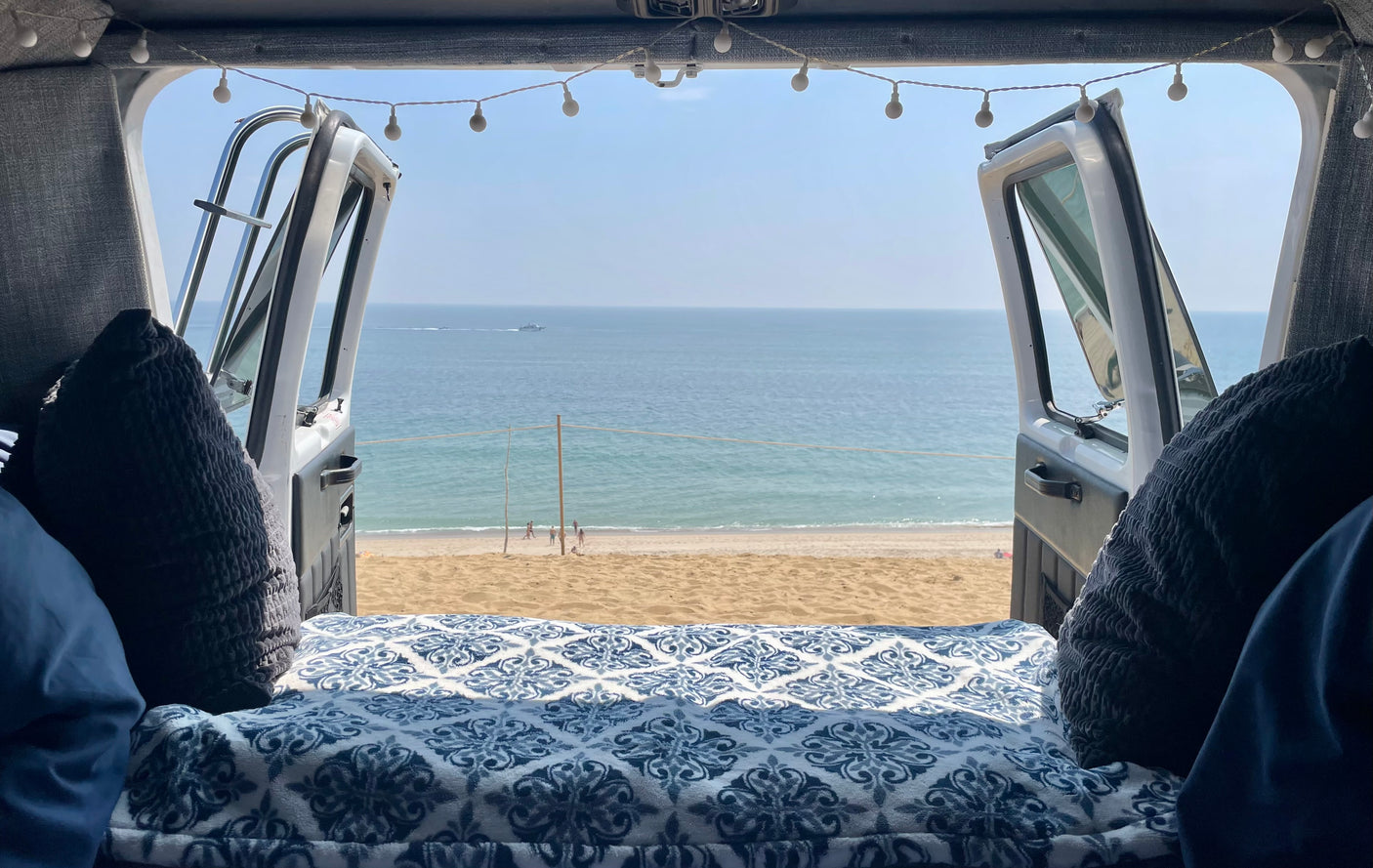 Relaxing **RV rental** view of a serene beach and ocean, with people and boats, from a cozy bed inside a camper van.