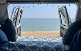 Relaxing **RV rental** view of a serene beach and ocean, with people and boats, from a cozy bed inside a camper van.