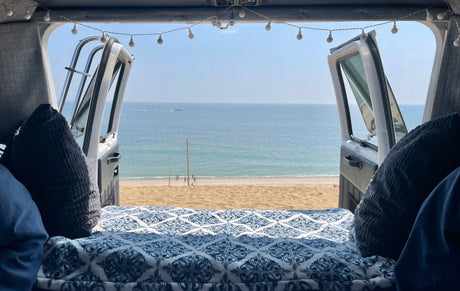 Relaxing **RV rental** view of a serene beach and ocean, with people and boats, from a cozy bed inside a camper van.