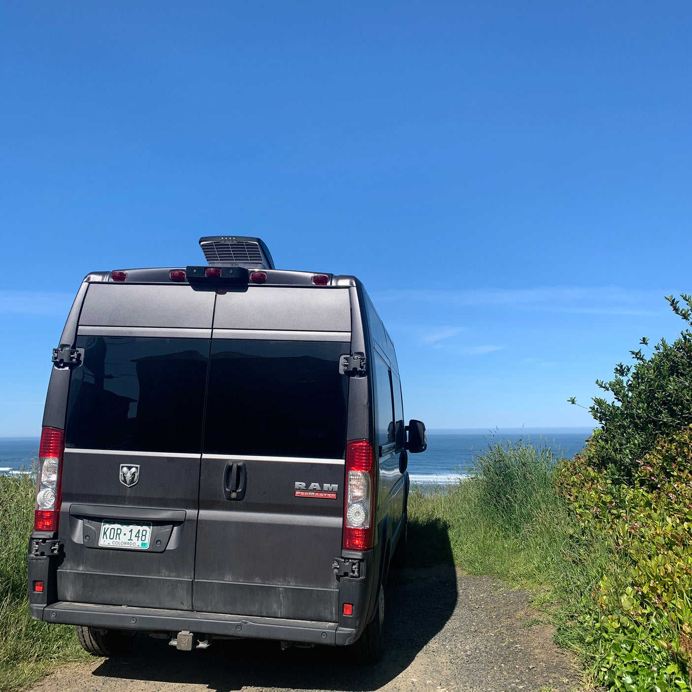 A grey RAM ProMaster RV rental van with a rooftop AC unit is parked overlooking a sunny ocean and green foliage.