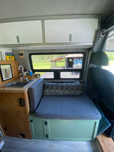 This RV rental interior shows a compact kitchen with a sink next to a bench seat. Overhead cabinets and a window overlook green scenery.