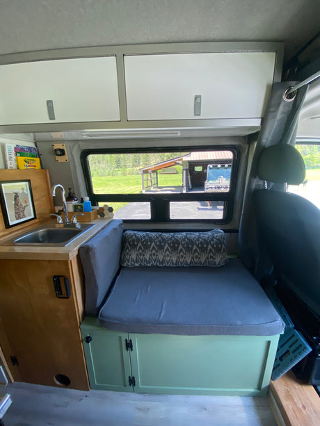 This RV rental interior shows a compact kitchen with a sink next to a bench seat. Overhead cabinets and a window overlook green scenery.