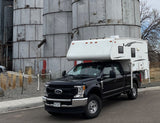 Black Ford Super Duty truck with truck camper, like an RV rental, parked by metal silos.