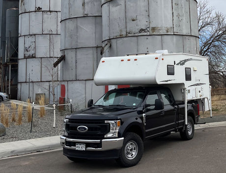 Black Ford Super Duty truck with truck camper, like an RV rental, parked by metal silos.