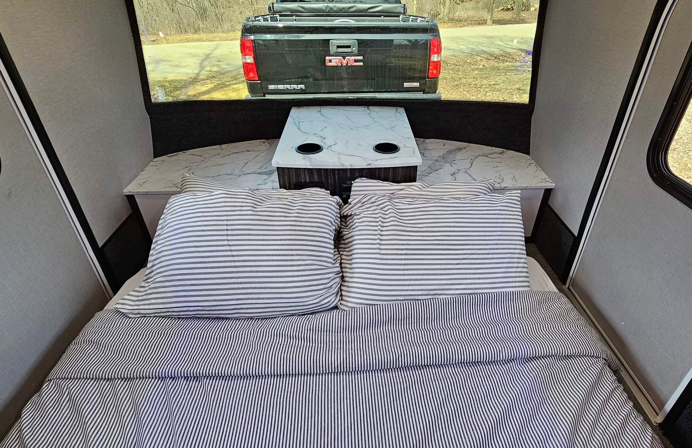 Cozy RV rental interior with striped bed, small table, and a view of a truck through the rear window.