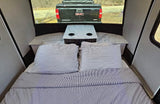 Cozy RV rental interior with striped bed, small table, and a view of a truck through the rear window.