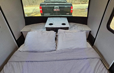 Cozy RV rental interior with striped bed, small table, and a view of a truck through the rear window.