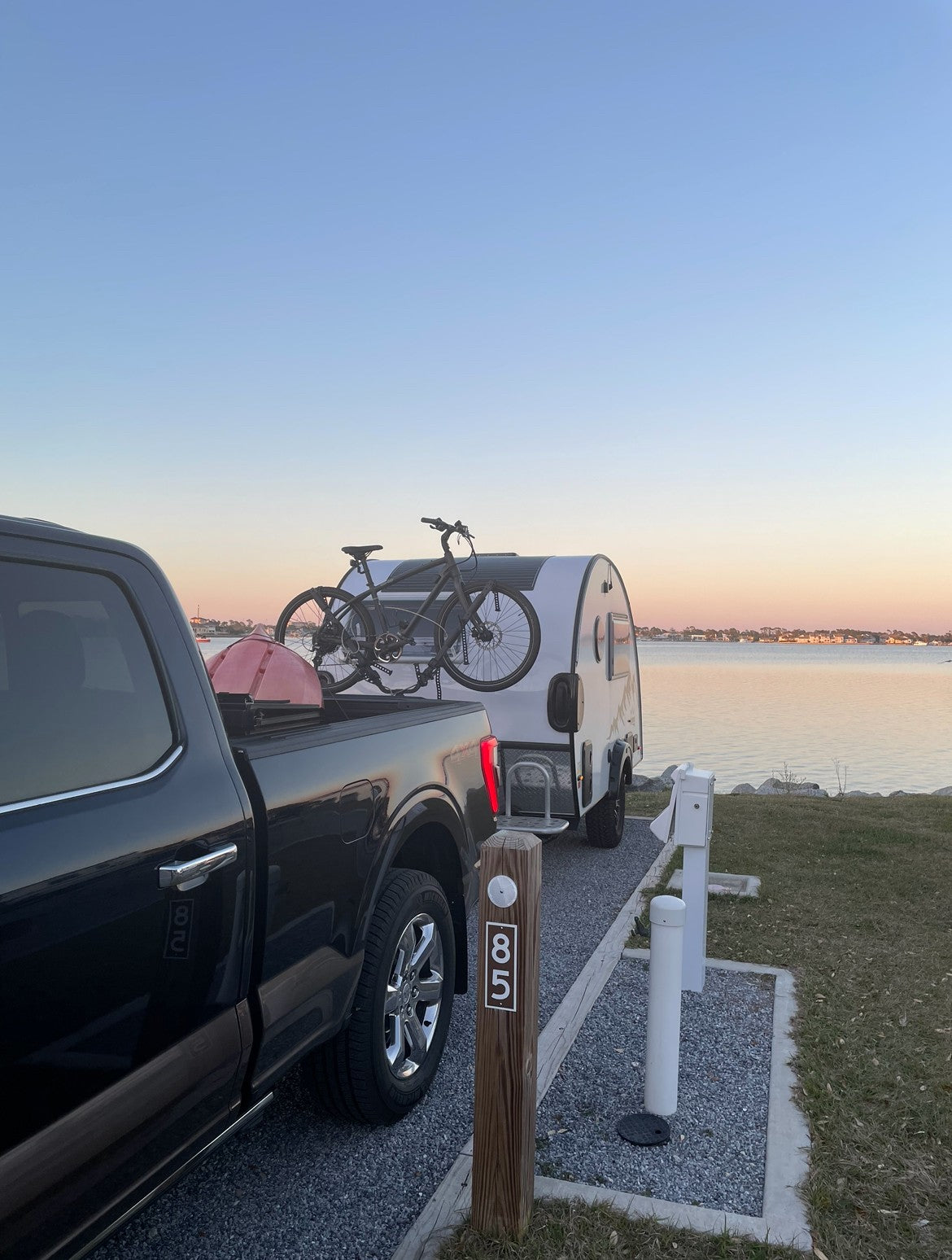 Campsite 85: A serene waterfront RV rental setup at dusk. Black truck tows a teardrop RV with bikes for waterside adventures.