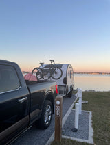 Campsite 85: A serene waterfront RV rental setup at dusk. Black truck tows a teardrop RV with bikes for waterside adventures.