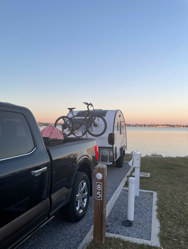 Campsite 85: A serene waterfront RV rental setup at dusk. Black truck tows a teardrop RV with bikes for waterside adventures.