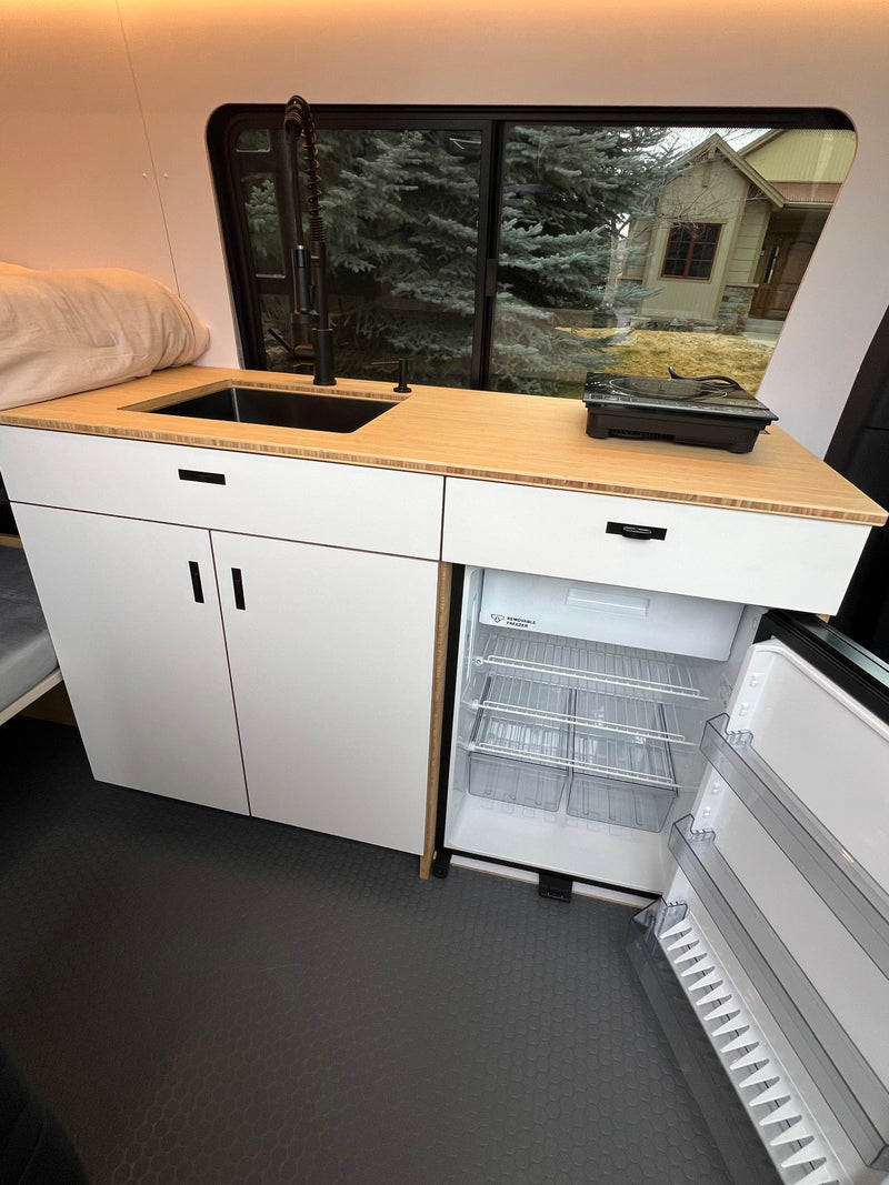 Modern RV rental kitchen with white cabinets, bamboo counter, sink, induction cooktop, and open refrigerator/freezer. — photo 14
