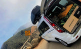 RV rental van with a Thule roof box and camper bed setup, parked on a scenic mountain road near a "steep slope" sign.