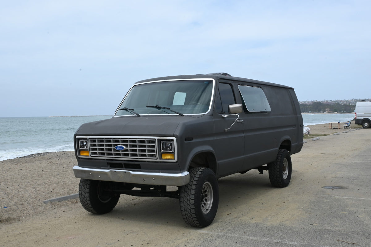 Adventure-ready dark gray Ford van parked by the ocean, ideal as an RV rental for beach trips.