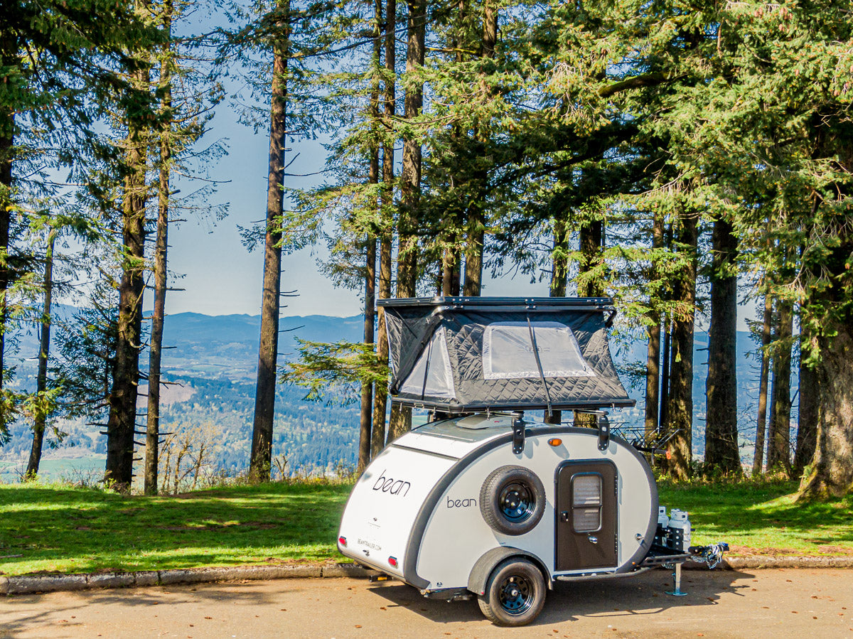 A white Bean teardrop trailer with a deployed rooftop tent, ideal for an RV rental. Parked at a scenic mountain overlook.