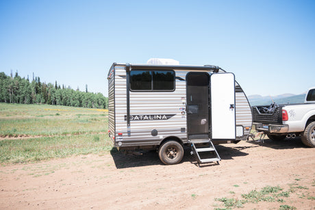 An RV rental with its door open, hitched to a pickup truck on a dirt road, amidst trees, fields, & distant mountains.