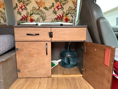 RV rental: Inside a campervan, a custom wood kitchenette with sink, water jug, storage, and floral curtains.