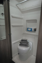 Compact RV rental wet bath featuring a toilet, shower, and built-in storage. Gray bags are stacked on the toilet lid.