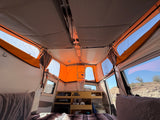 Inside a pop-up rv rental: compact interior with bed, small kitchen, and panoramic windows showing a desert view.